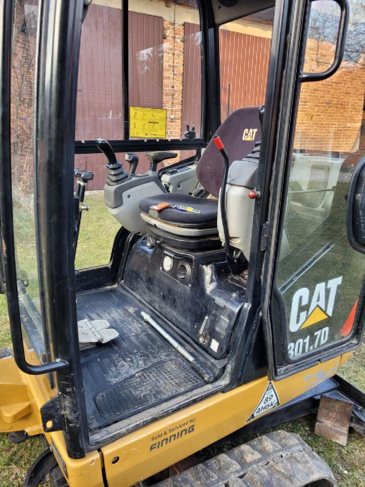 Wnętrze kabiny mini koparki CAT 301.7D, widoczne siedzenie operatora z narzędziami, dźwignie sterujące i naklejka informacyjna na szybie, z zewnątrz logo CAT i oznaczenia modelu.