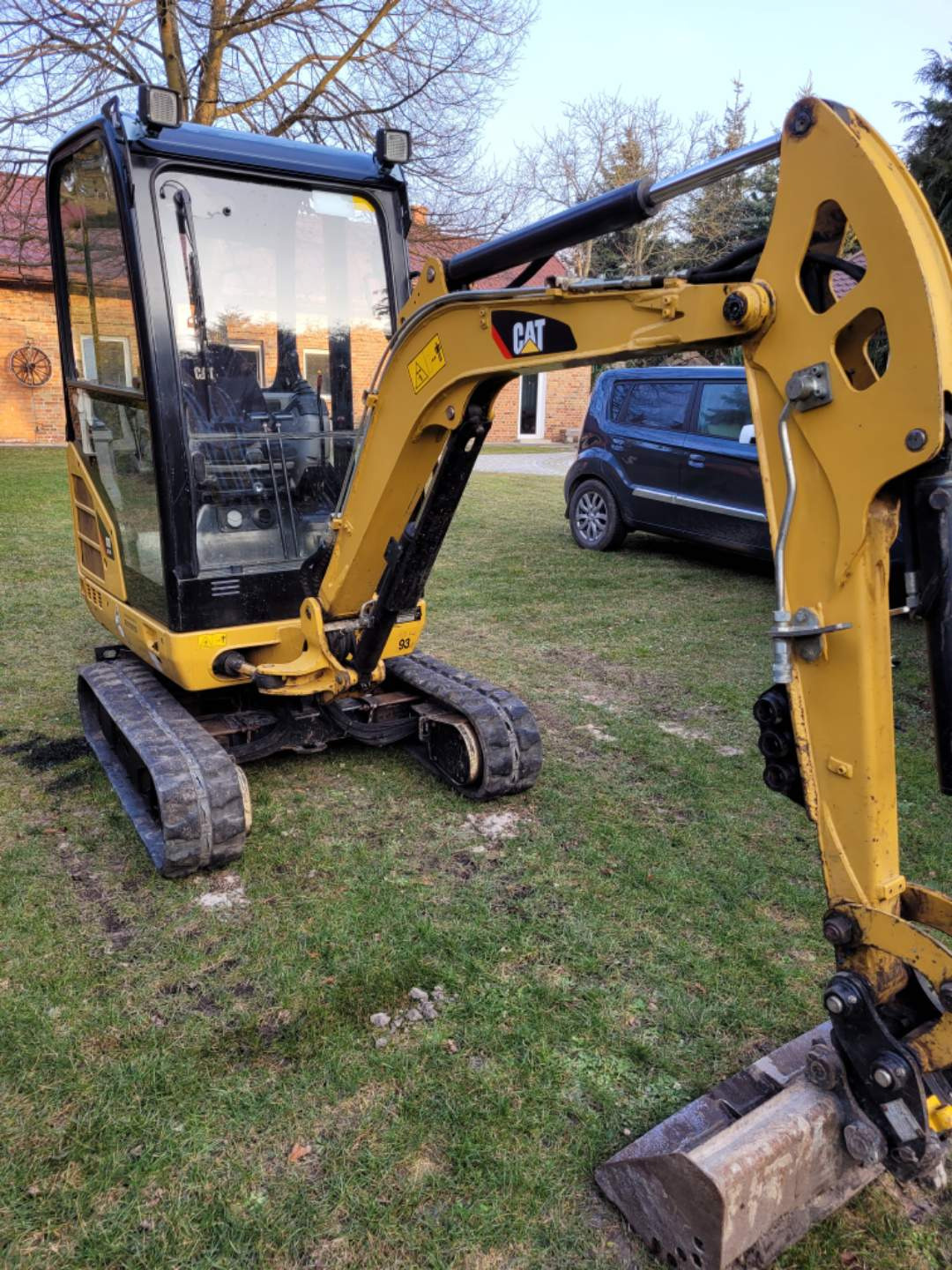 Żółta mini koparka CAT na trawiastym terenie, z widocznym domem z czerwonej cegły i ciemnym samochodem osobowym w tle.