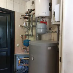 Instalacja centralnego ogrzewania: zbiornik akumulacyjny, zasobnik ciepłej wody użytkowej, naczynia wzbiorcze i rozdzielacz z pompą obiegową w kotłowni z wykafelkowanymi ścianami.