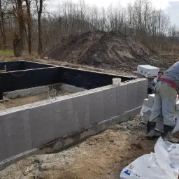 Budowa fundamentów: pracownik w zielonym kasku układa bloczki fundamentowe, widoczna izolacja pionowa, w tle sterta ziemi i budynek mieszkalny.