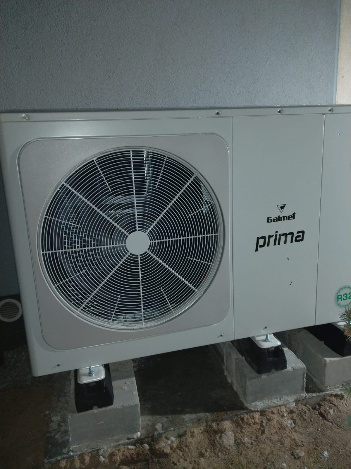 Zewnętrzna jednostka pompy ciepła Galmet Prima, zamontowana na betonowych bloczkach z gumowymi podkładkami antywibracyjnymi, widoczna szara ściana budynku w tle.