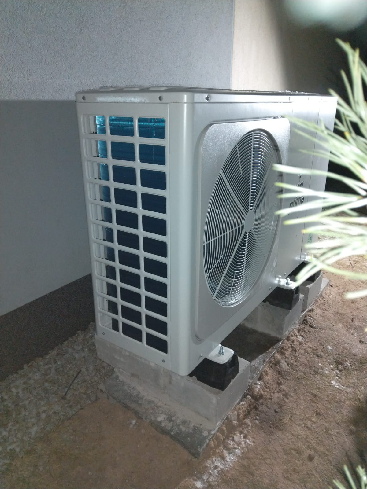 Zewnętrzna jednostka klimatyzacji marki Prima, zamontowana na betonowych bloczkach z gumowymi podkładkami, widoczna krata wlotowa i wentylator.