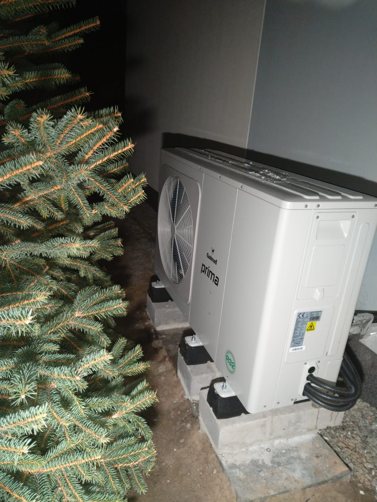 Zewnętrzna jednostka klimatyzacji Galmet Prima, zamontowana na betonowych podstawach obok iglastego drzewka, z widocznymi przewodami i tabliczką znamionową.