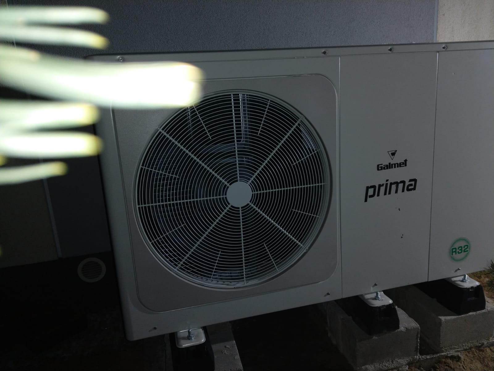 Szara pompa ciepła Galmet Prima R32 zamontowana na betonowych bloczkach, widoczna z boku z fragmentem roślinności w lewym górnym rogu.