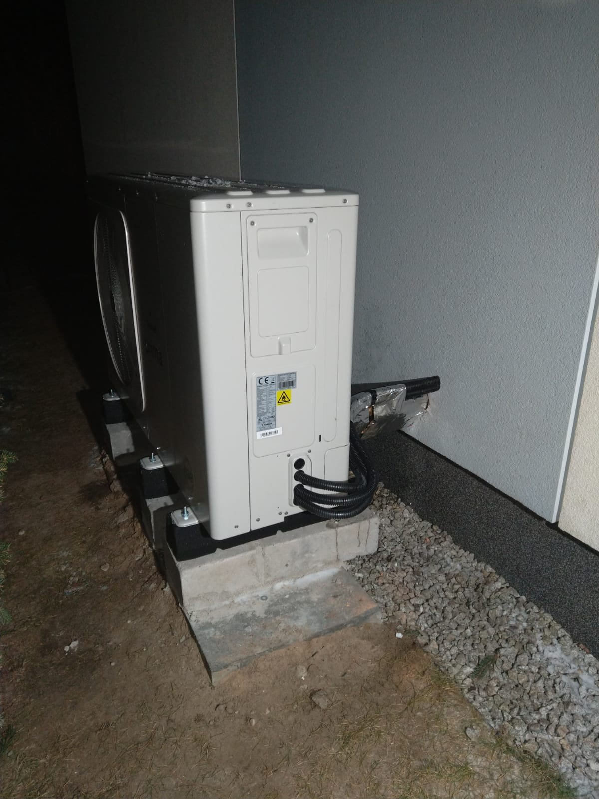 Zewnętrzna jednostka klimatyzacji zamontowana na betonowej podstawie z gumowymi amortyzatorami, rury odprowadzające wilgoć zabezpieczone folią aluminiową.