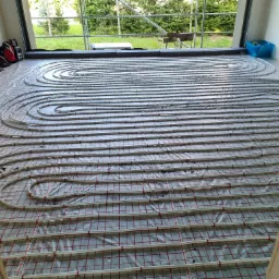 Montaż ogrzewania podłogowego w nowym budynku: rury grzewcze ułożone w pętlach na folii z siatką, widoczne narzędzia i torba, okno z widokiem na zewnątrz.