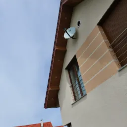 Fragment elewacji domu z beżowym tynkiem, dekoracyjnymi pasami w kolorze piaskowym, brązową stolarką okienną i satelitarną anteną na ścianie, widok z dołu.