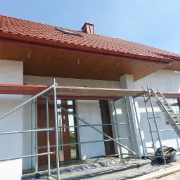 Dom jednorodzinny w trakcie ocieplania elewacji, widoczne rusztowania, zabezpieczona folią podstawa, dach z brązowej dachówki, okno dachowe i komin.