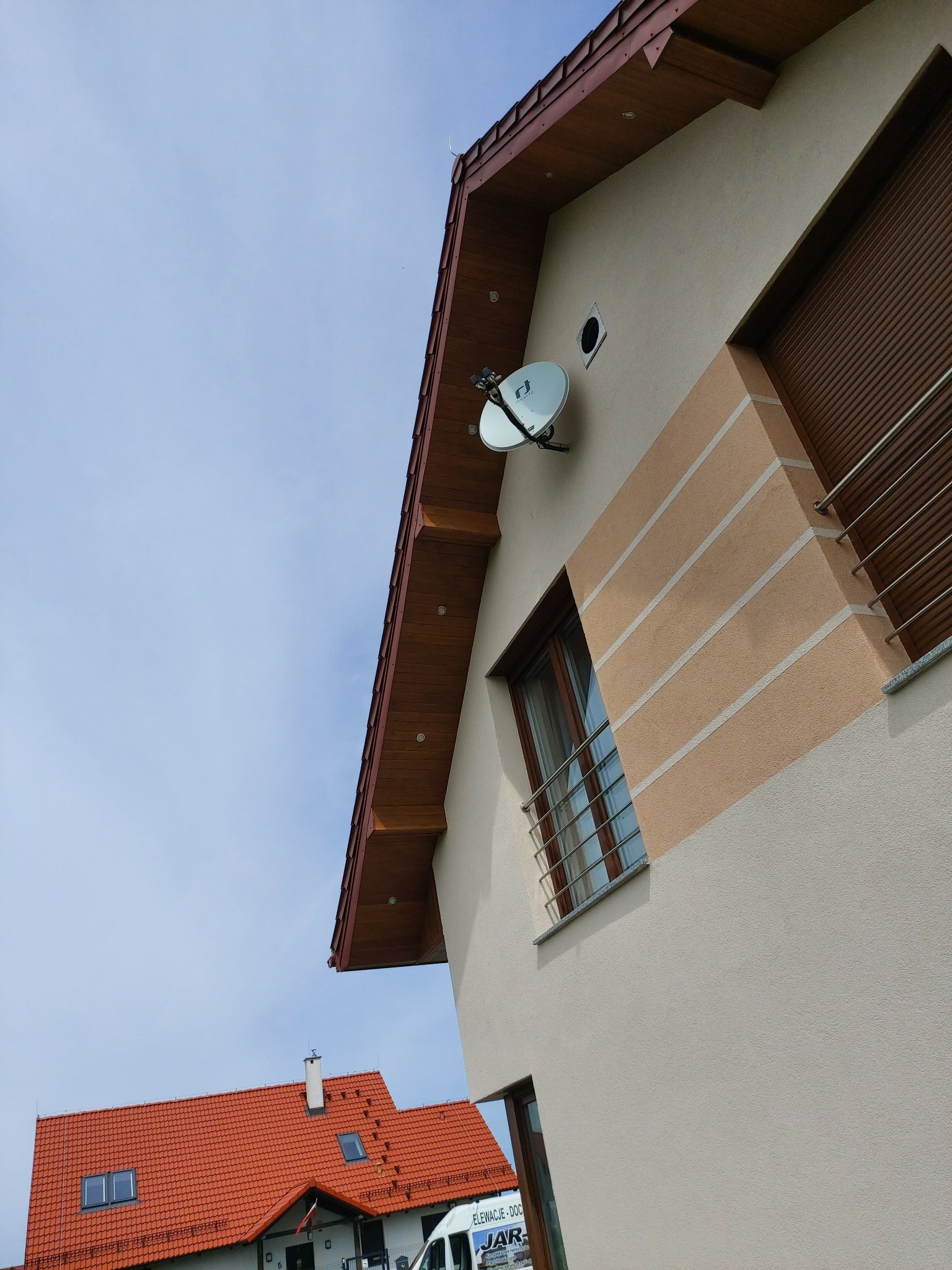 Fragment elewacji domu z beżowym tynkiem, dekoracyjnymi pasami w kolorze piaskowym, brązową stolarką okienną i satelitarną anteną na ścianie, widok z dołu.