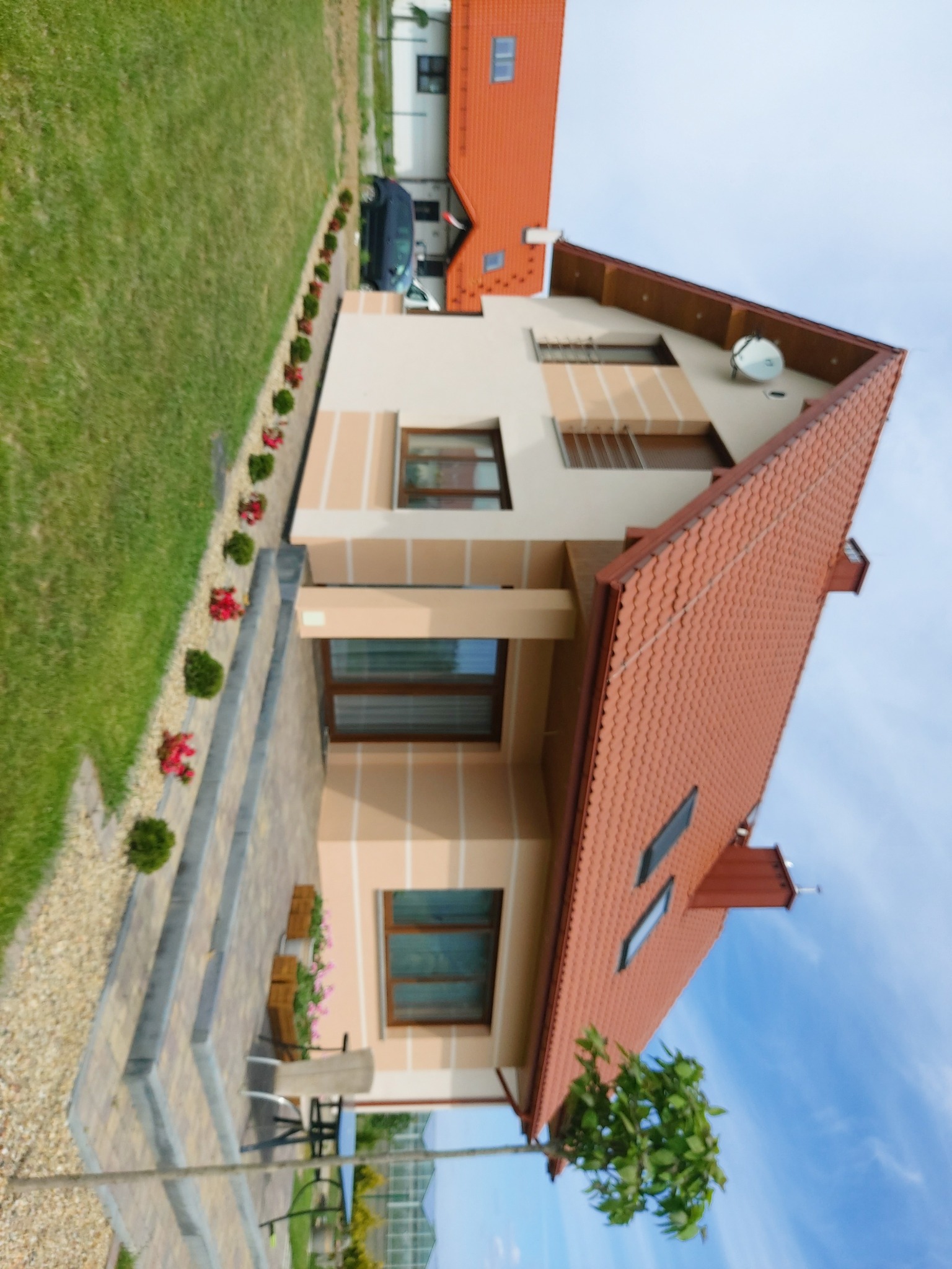 Widok z boku na dom jednorodzinny z beżową elewacją, brązowymi oknami i czerwoną dachówką, z zadbanym trawnikiem, ozdobnymi krzewami i kwiatami wzdłuż ścieżki oraz meblami ogrodowymi na tarasie.