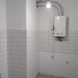 Łazienka w trakcie remontu z ułożonymi białymi, prostokątnymi płytkami, zamontowanym piecykiem gazowym Daikin i widocznymi narzędziami na podłodze z paneli.