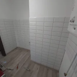 Świeżo wykafelkowana łazienka z białymi, prostokątnymi płytkami, drewnopodobna podłoga, widoczne narzędzia glazurnicze i piec gazowy na ścianie.