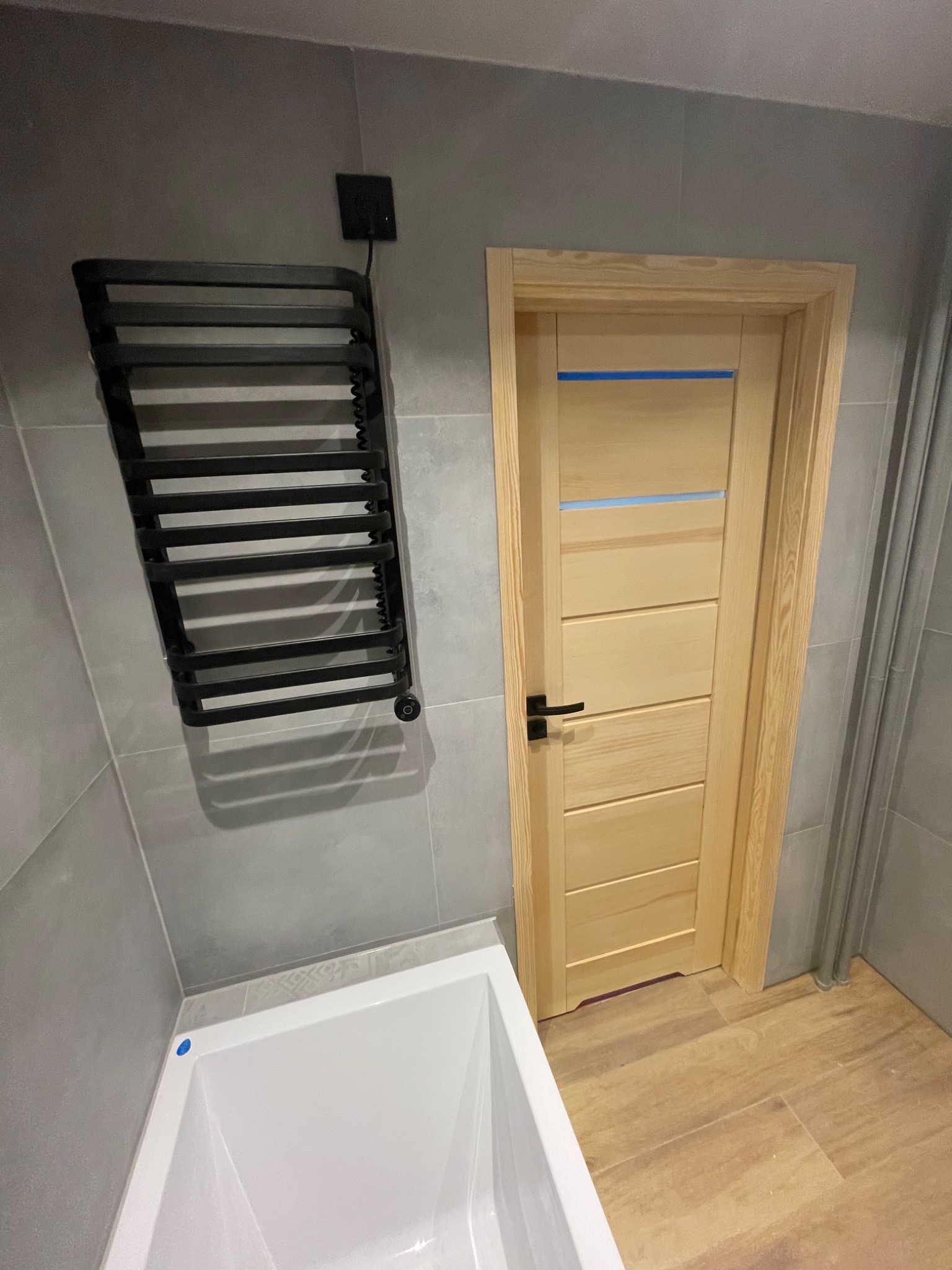 Wykończona łazienka z drewnianą ramą drzwi, czarnym grzejnikiem drabinkowym i białą wanną, płytki w odcieniach szarości.