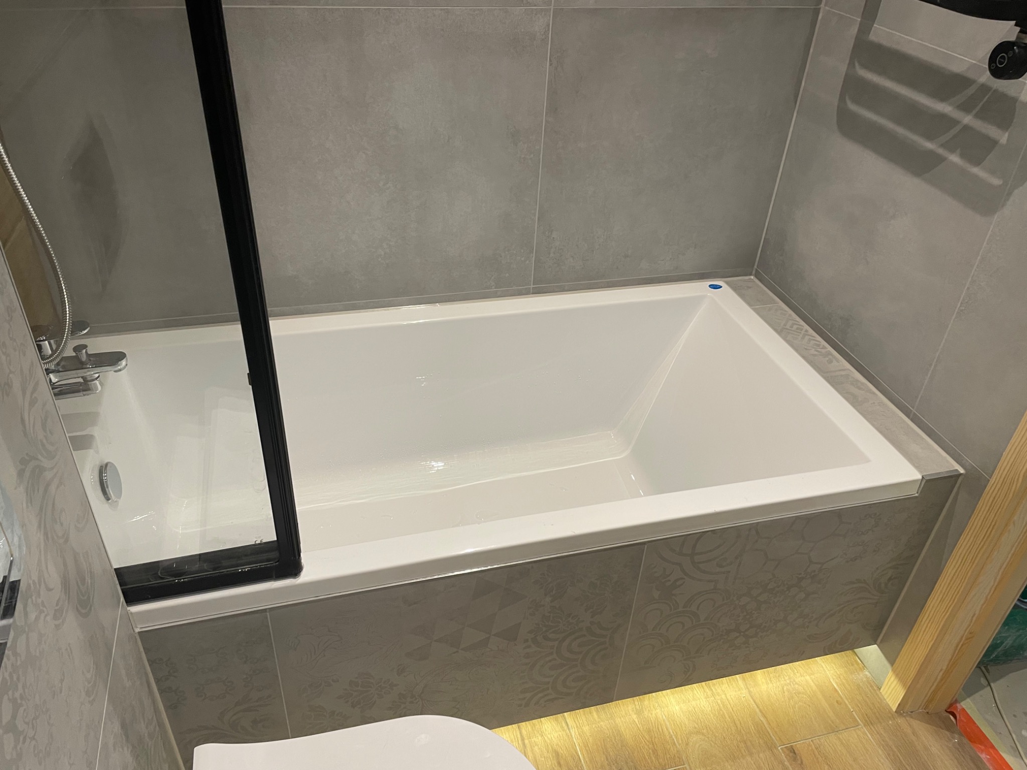 Biała wanna z czarną ramą kabiny prysznicowej, obłożona szarymi płytkami z wzorem, podświetlona od dołu listwą LED, szare płytki na ścianie, widoczny fragment toalety i drewnianej ramy.