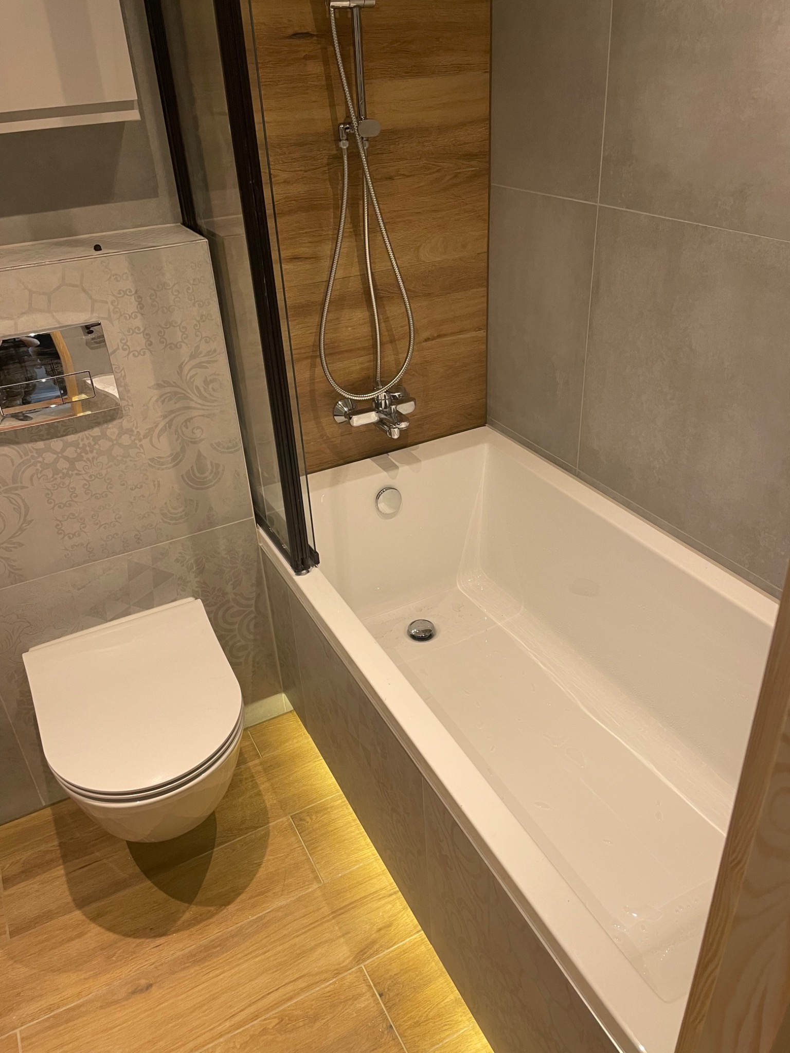 Nowoczesna łazienka z wanną z parawanem, wiszącą toaletą i drewnopodobnymi płytkami podłogowymi z delikatnym podświetleniem LED.