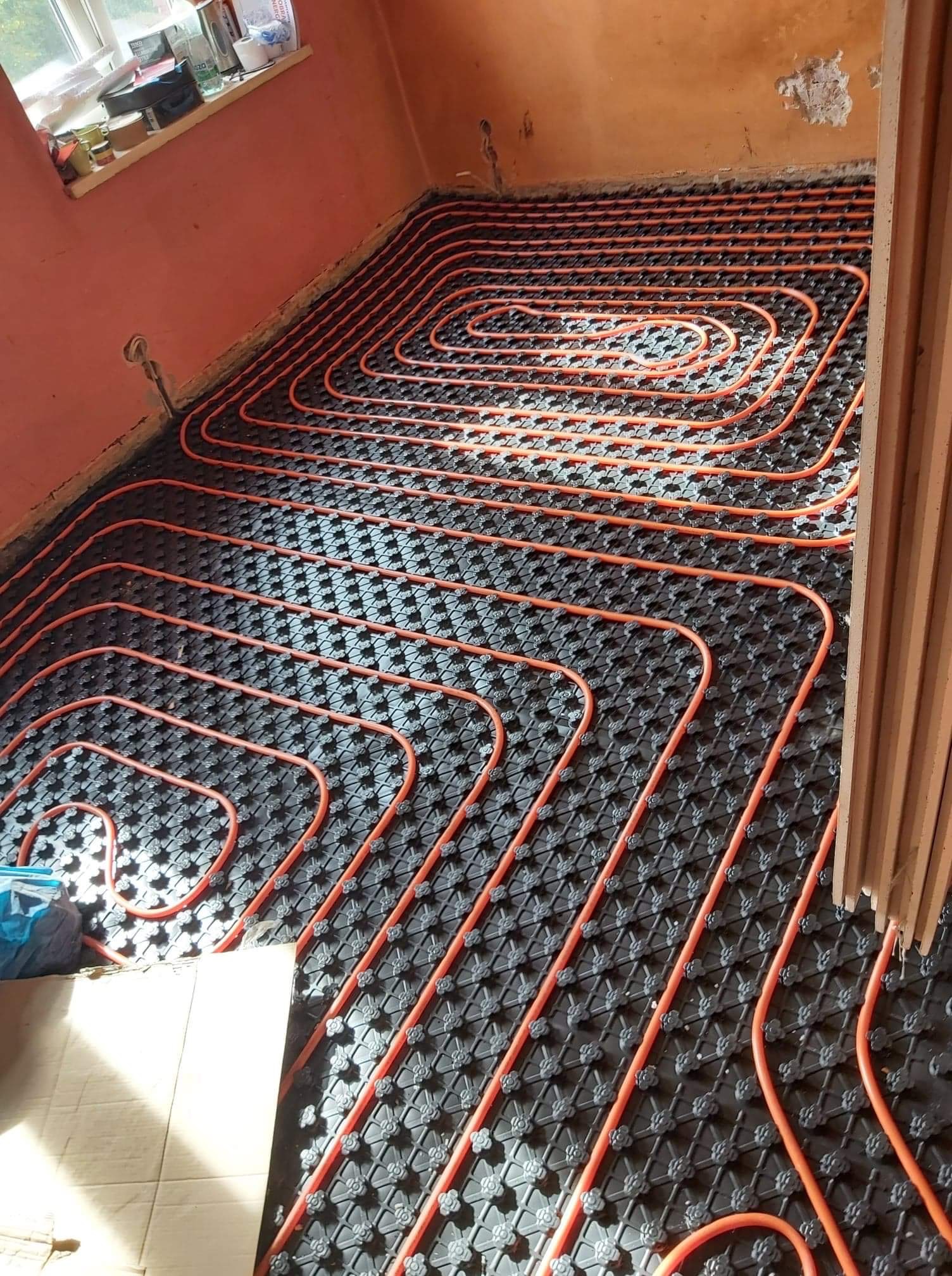 Pomarańczowe rury ogrzewania podłogowego ułożone spiralnie na czarnej macie z wypustkami, widoczne niedoskonałości ściany i fragment okna z parapetem.