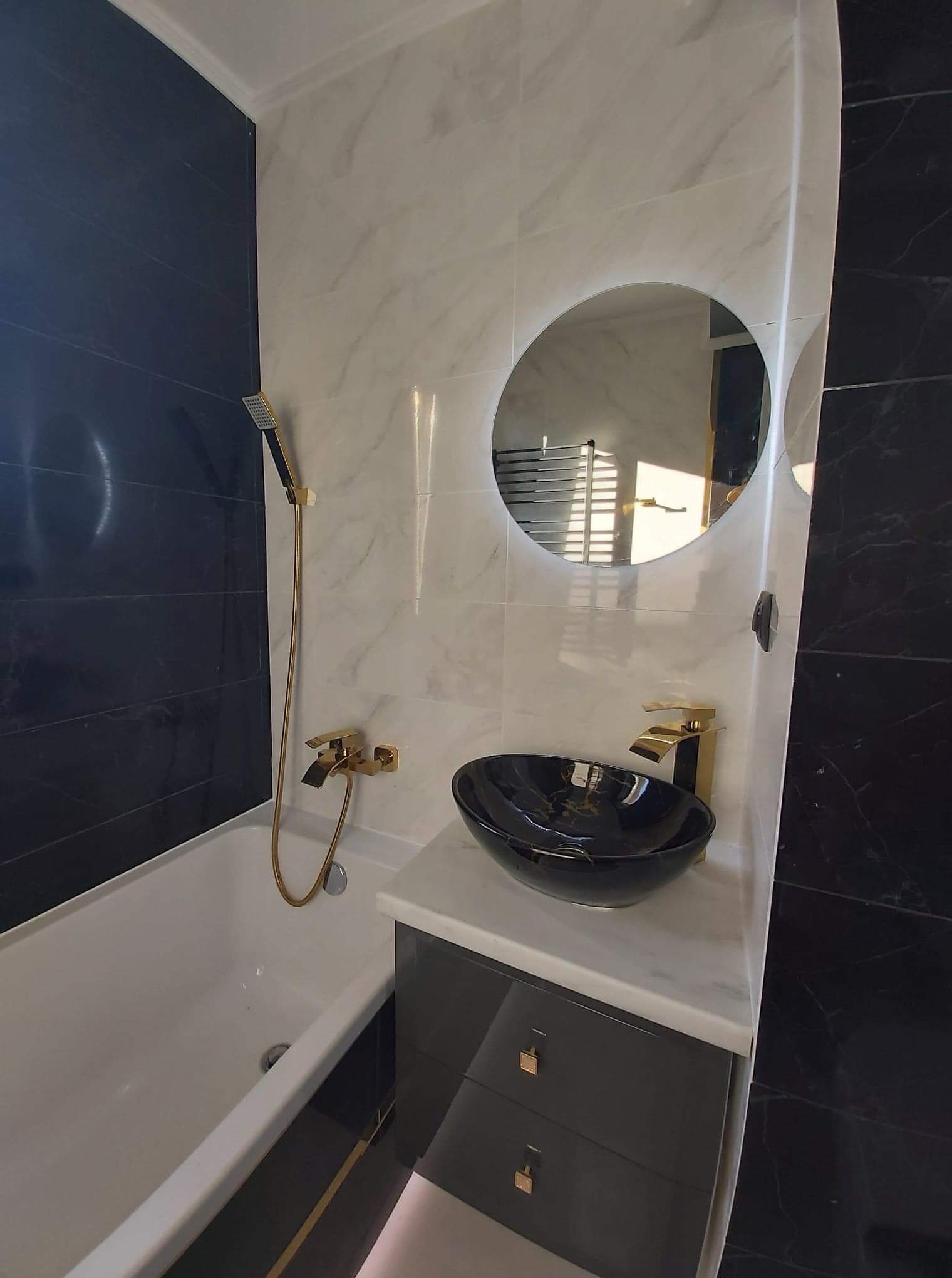 Nowoczesna łazienka z czarno-białymi marmurowymi płytkami, złotą armaturą i okrągłym lustrem z podświetleniem LED odbijającym grzejnik.