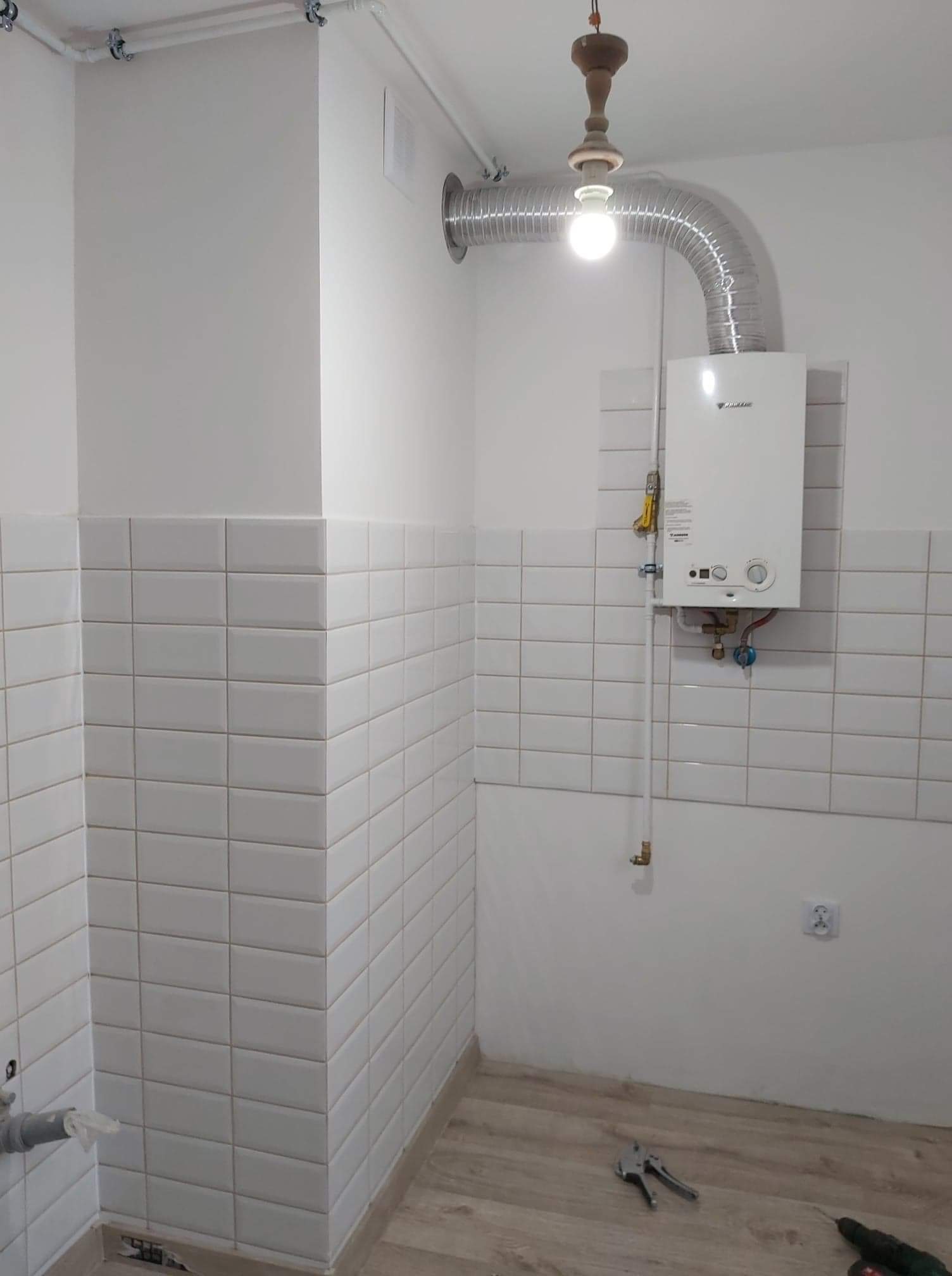 Łazienka w trakcie remontu z ułożonymi białymi, prostokątnymi płytkami, zamontowanym piecykiem gazowym Daikin i widocznymi narzędziami na podłodze z paneli.