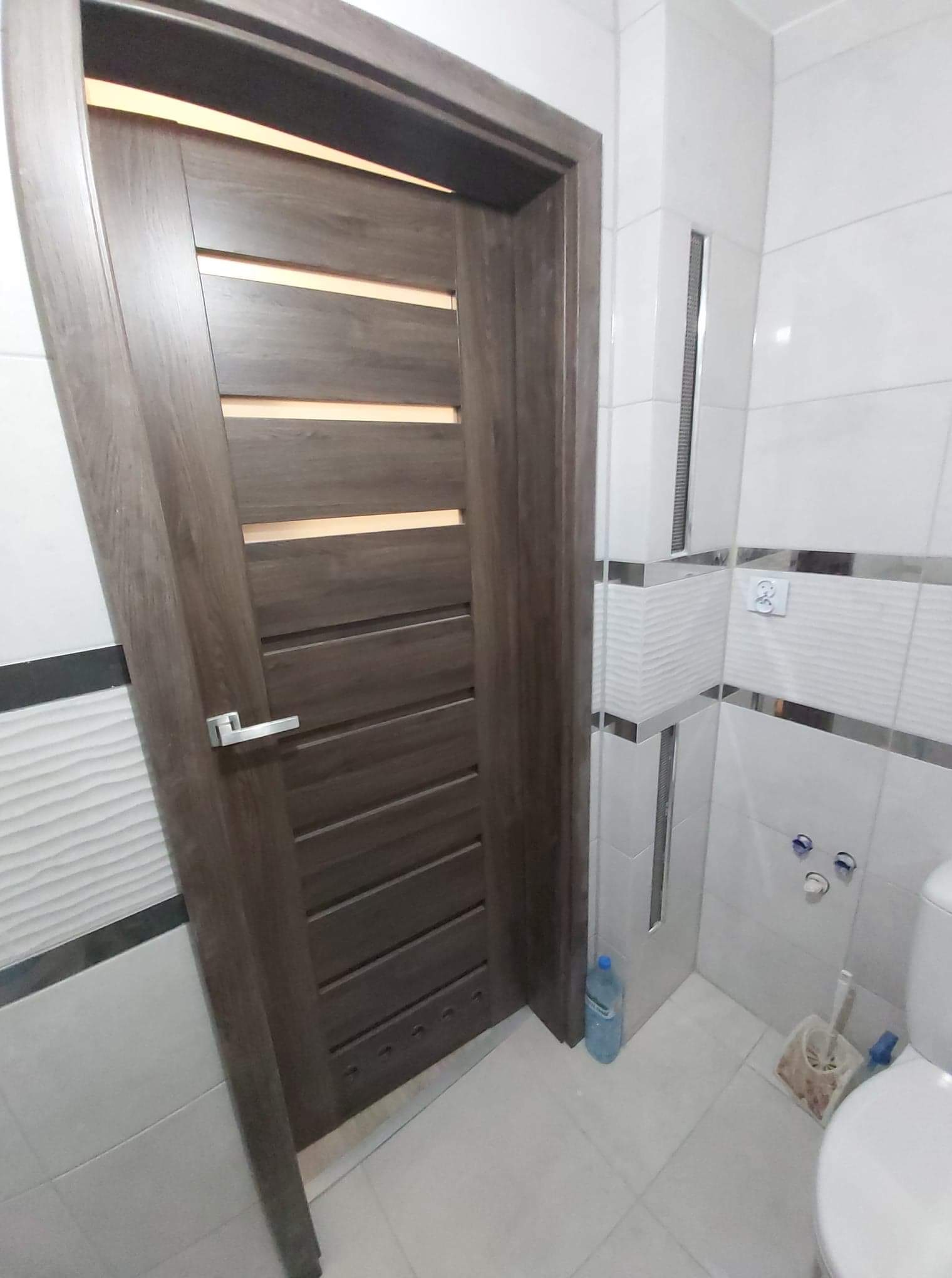 Nowoczesna łazienka z drewnianymi drzwiami z poziomymi przeszkleniami, białe płytki z dekoracyjnymi paskami, widoczne elementy toalety i butelka z wodą na podłodze.