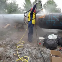 Pracownik w kombinezonie ochronnym piaskuje fragment rurociągu na zewnątrz, otoczony pyłem i sprzętem.