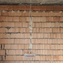 Wewnętrzna ściana z czerwonej cegły z zamontowanymi przewodami elektrycznymi i puszkami, zabezpieczonymi gipsem, widoczne schody w surowym stanie i okno z pianką montażową.