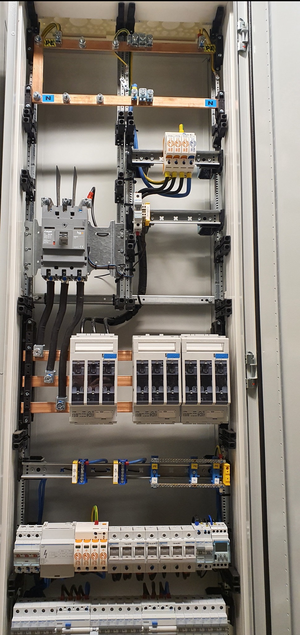 Wewnętrzny widok rozdzielnicy elektrycznej z szynami miedzianymi, bezpiecznikami i zabezpieczeniami różnicowoprądowymi, okablowanie w peszlach, widoczne oznaczenia N i PE.