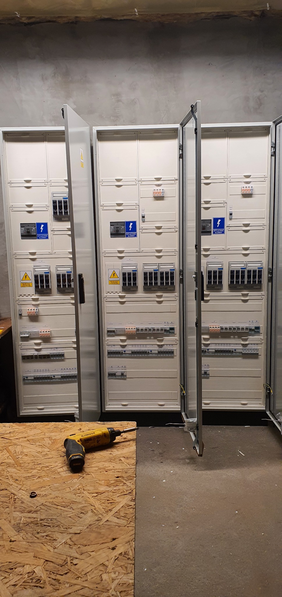 Rozdzielnice elektryczne w szarych obudowach z otwartymi drzwiami, widoczne bezpieczniki i oznaczenia, na pierwszym planie wkrętarka DeWalt na płycie OSB.