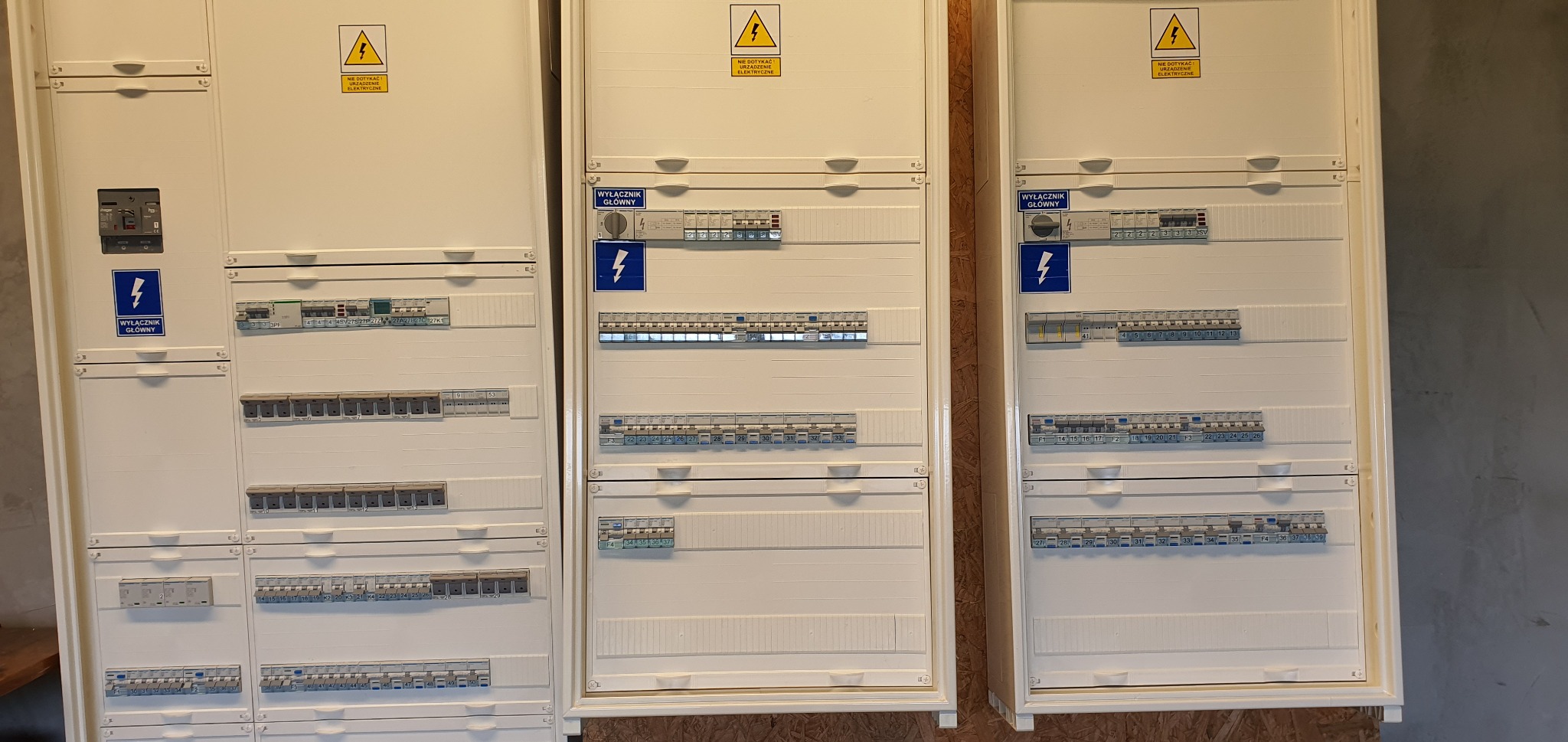 Trzy białe skrzynki rozdzielcze z bezpiecznikami i wyłącznikami, widoczne żółte ostrzeżenia o napięciu, etykiety z opisami obwodów.