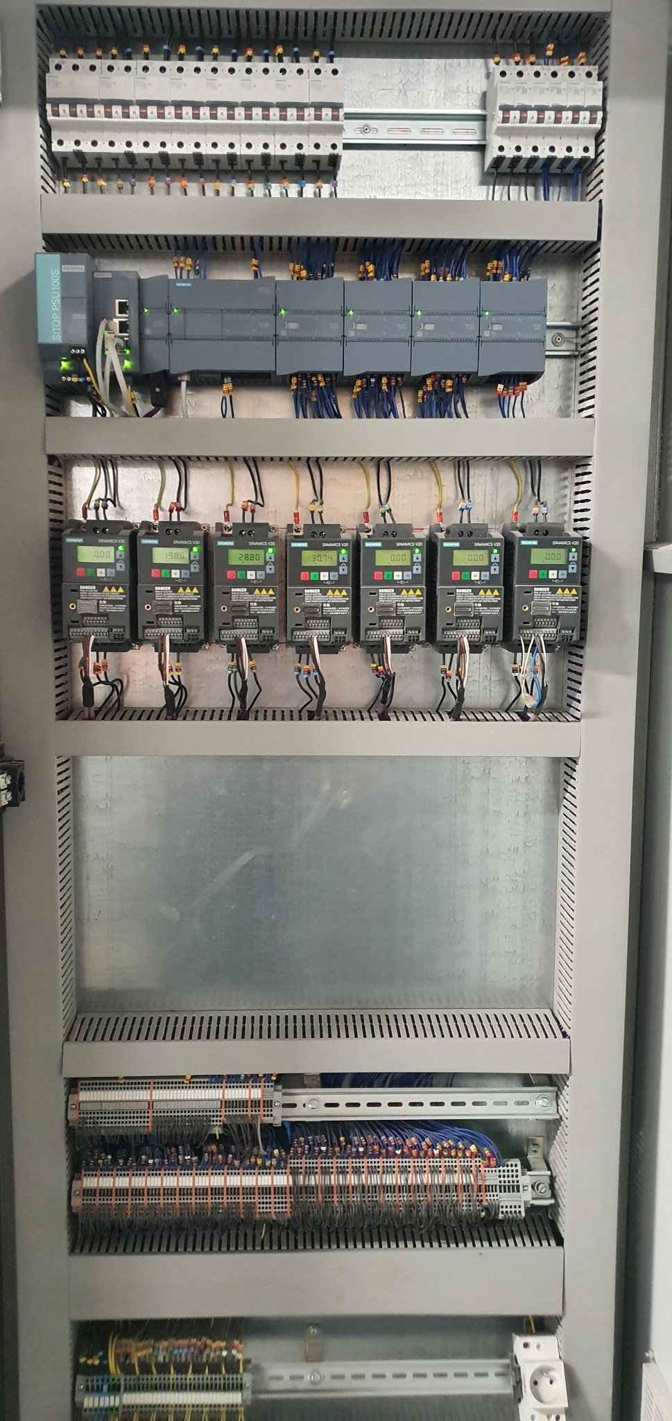 Rozdzielnica elektryczna z zamontowanymi modułami sterującymi Siemens Sinamics V20 z wyświetlaczami pokazującymi różne wartości, widoczne okablowanie i bezpieczniki, całość w metalowej obudowie.