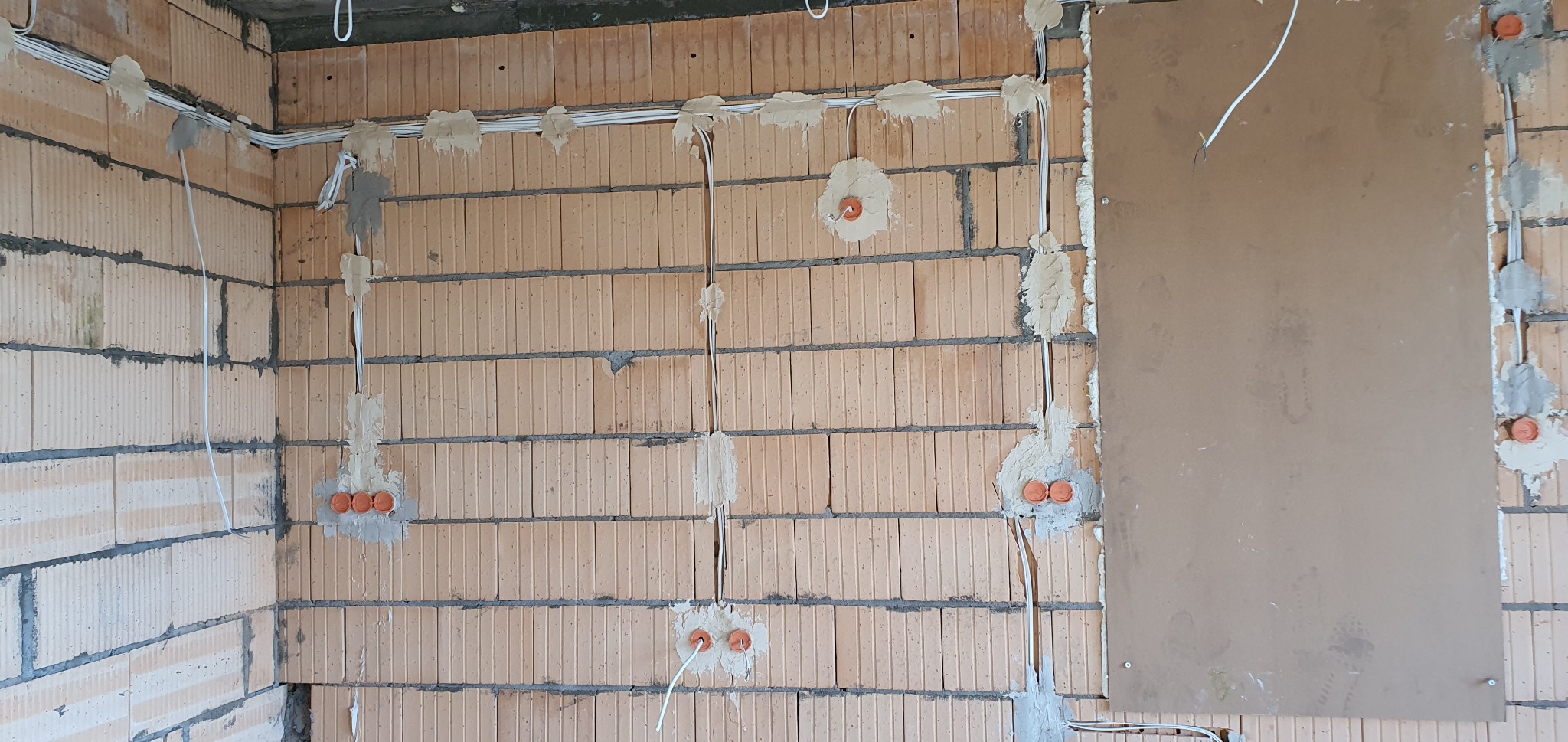 Instalacja elektryczna w trakcie montażu w nowym budynku w Zbąszyniu. Widoczne przewody rozprowadzone po ścianach z cegły, zamocowane gipsem, puszki instalacyjne w kolorze pomarańczowym.