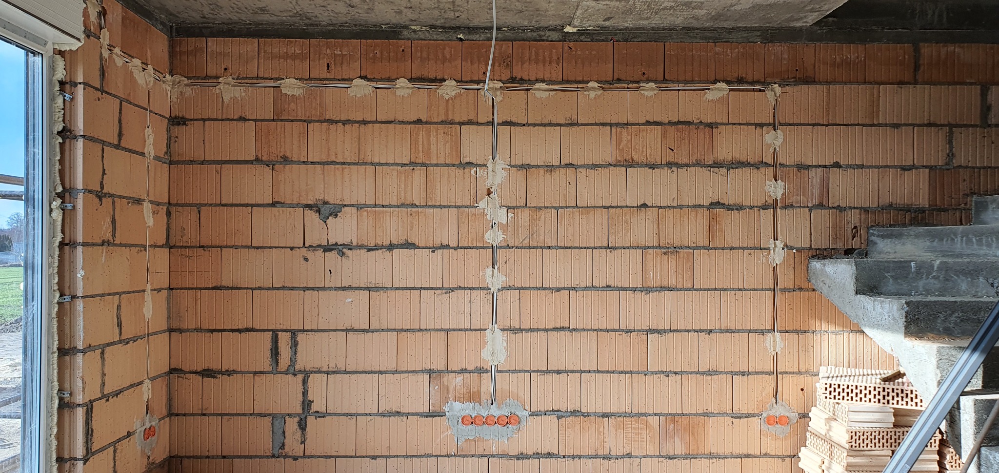 Wewnętrzna ściana z czerwonej cegły z zamontowanymi przewodami elektrycznymi i puszkami, zabezpieczonymi gipsem, widoczne schody w surowym stanie i okno z pianką montażową.