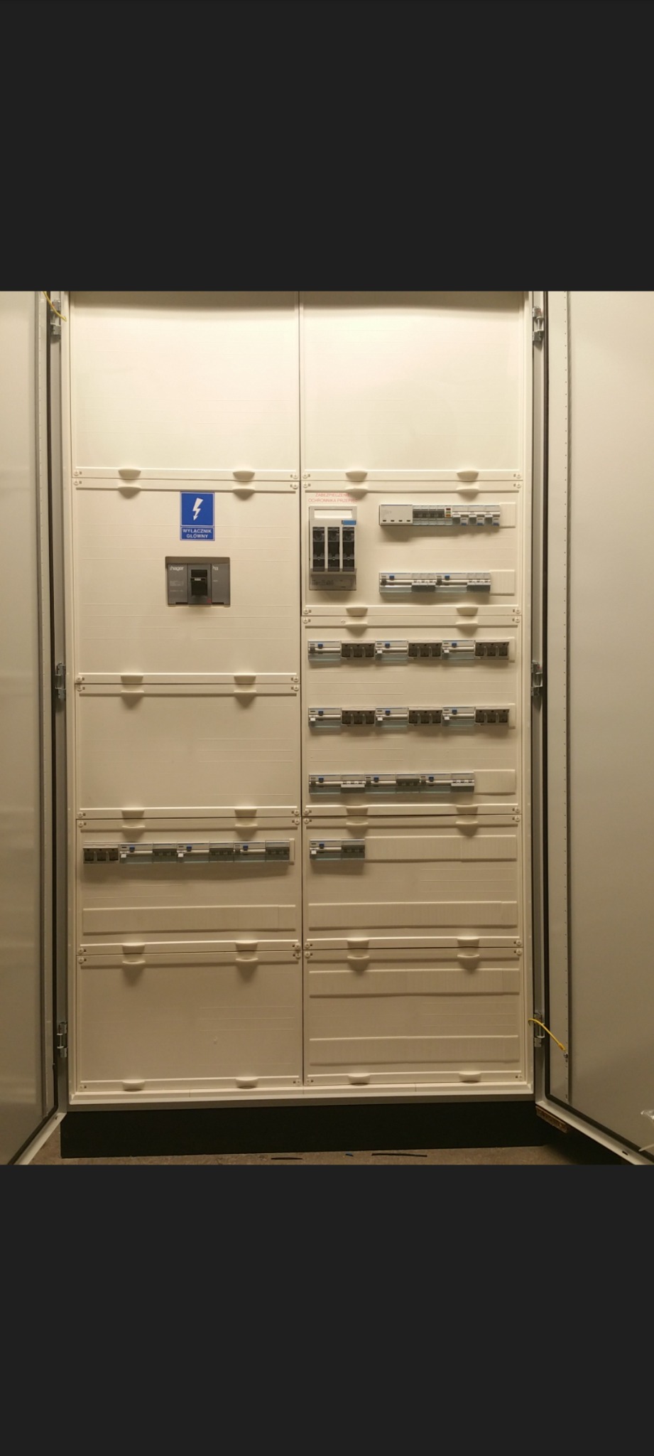 Otwarta, biała szafa rozdzielcza elektryczna z widocznymi bezpiecznikami, wyłącznikiem głównym i oznaczeniami, zamontowana w pomieszczeniu.