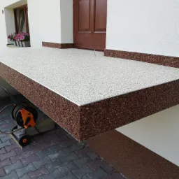 Wykończone schody wejściowe z brązowo-białą posadzką kamienną, widoczna zwinięta lanca do mycia ciśnieniowego i kostka brukowa na podjeździe.