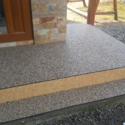 Wykończone schody wejściowe z posadzką żywiczną w kolorze szaro-beżowym, z wyraźnie oddzielonym stopniem w odcieniu piaskowym, przy wejściu do budynku z okładziną z kamienia naturalnego.