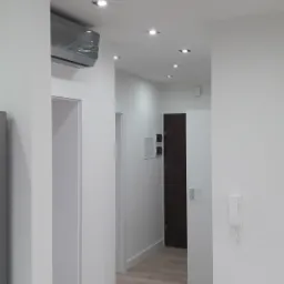 Wnętrze korytarza po remoncie, z jasnymi ścianami, panelami podłogowymi w odcieniach beżu i oświetleniem punktowym w suficie. Widoczny klimatyzator w folii ochronnej, białe futryny drzwiowe i biały...