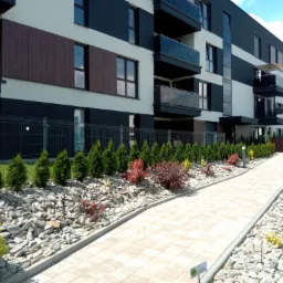 Nowoczesny budynek wielorodzinny z balkonami i zagospodarowanym terenem zielonym: ścieżka z kostki brukowej, rabata z kamieni, tuje i krzewy ozdobne wzdłuż ogrodzenia z siatki.