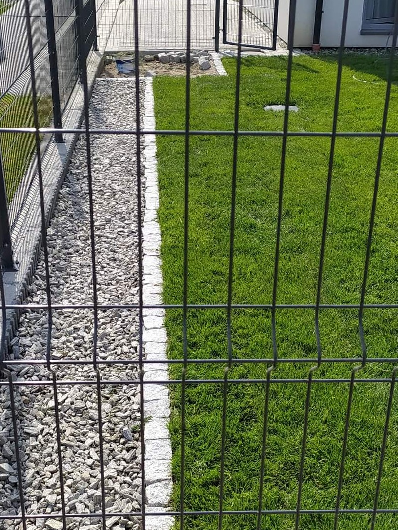 Panelowe ogrodzenie z podmurówką prefabrykowaną, obok wysypany pas żwiru i trawnik z obrzeżem z kostki granitowej, brama w tle.