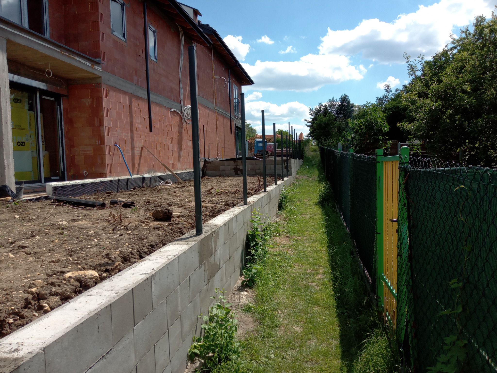 Wykop pod fundament ogrodzenia z betonowych bloczków, widoczne metalowe słupki, w tle budynek w trakcie budowy i siatka ogrodzeniowa z zieloną bramką.