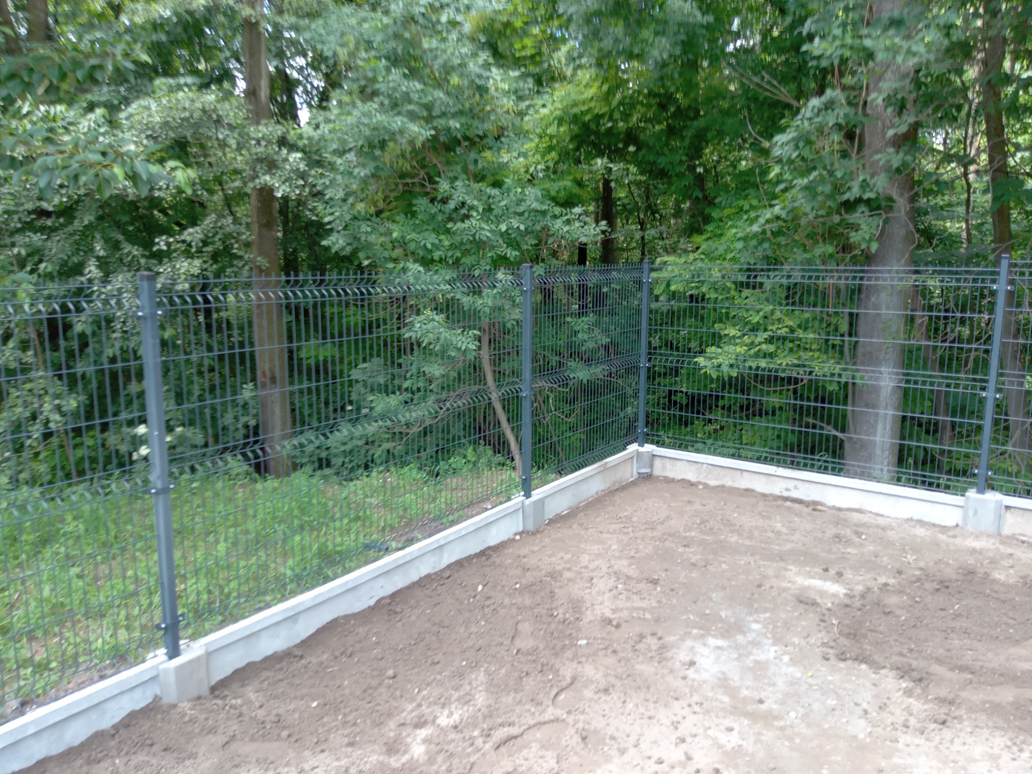 Nowo zainstalowane, szare ogrodzenie panelowe z betonową podmurówką na nierównym terenie, z widocznymi drzewami w tle.