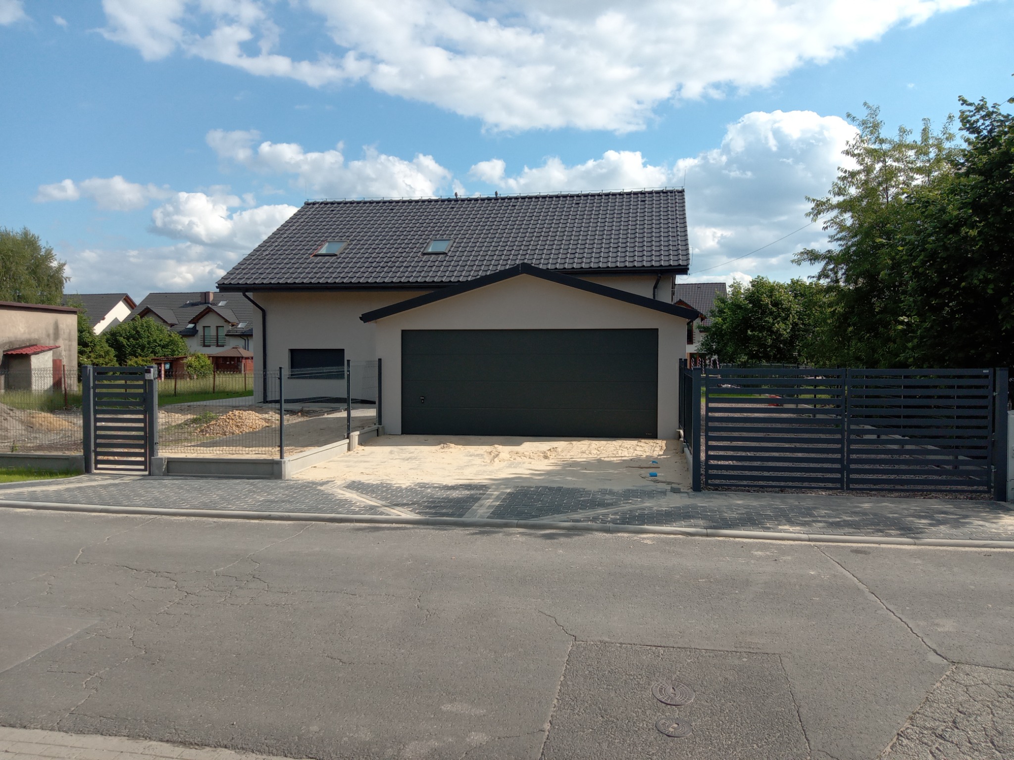 Nowoczesny dom jednorodzinny z grafitową bramą garażową i metalowym ogrodzeniem panelowym w kolorze antracytowym, widok z ulicy, kostka brukowa częściowo zasypana piaskiem.