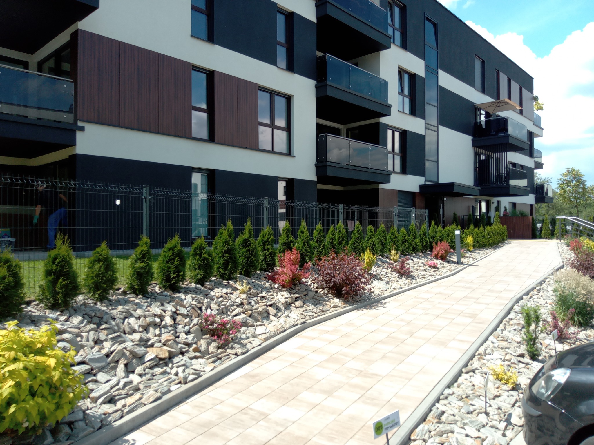 Nowoczesny budynek wielorodzinny z balkonami i zagospodarowanym terenem zielonym: ścieżka z kostki brukowej, rabata z kamieni, tuje i krzewy ozdobne wzdłuż ogrodzenia z siatki.