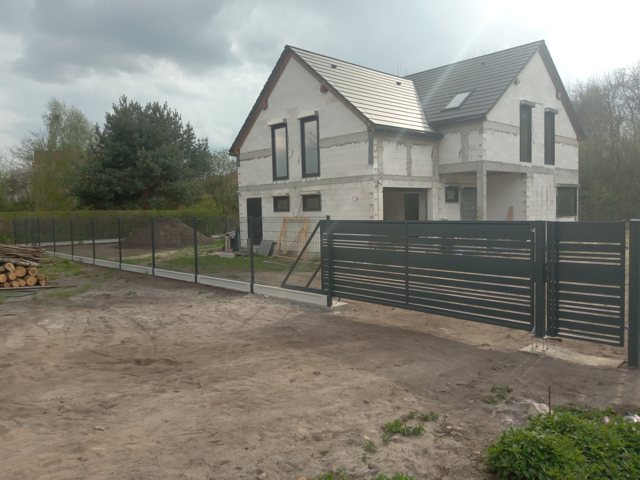 Nowoczesna, pozioma brama przesuwna w kolorze antracytowym, z widoczną konstrukcją mocującą, na tle surowego stanu domu z białego bloczka i ogrodzenia z siatki, stos drewna po lewej stronie.