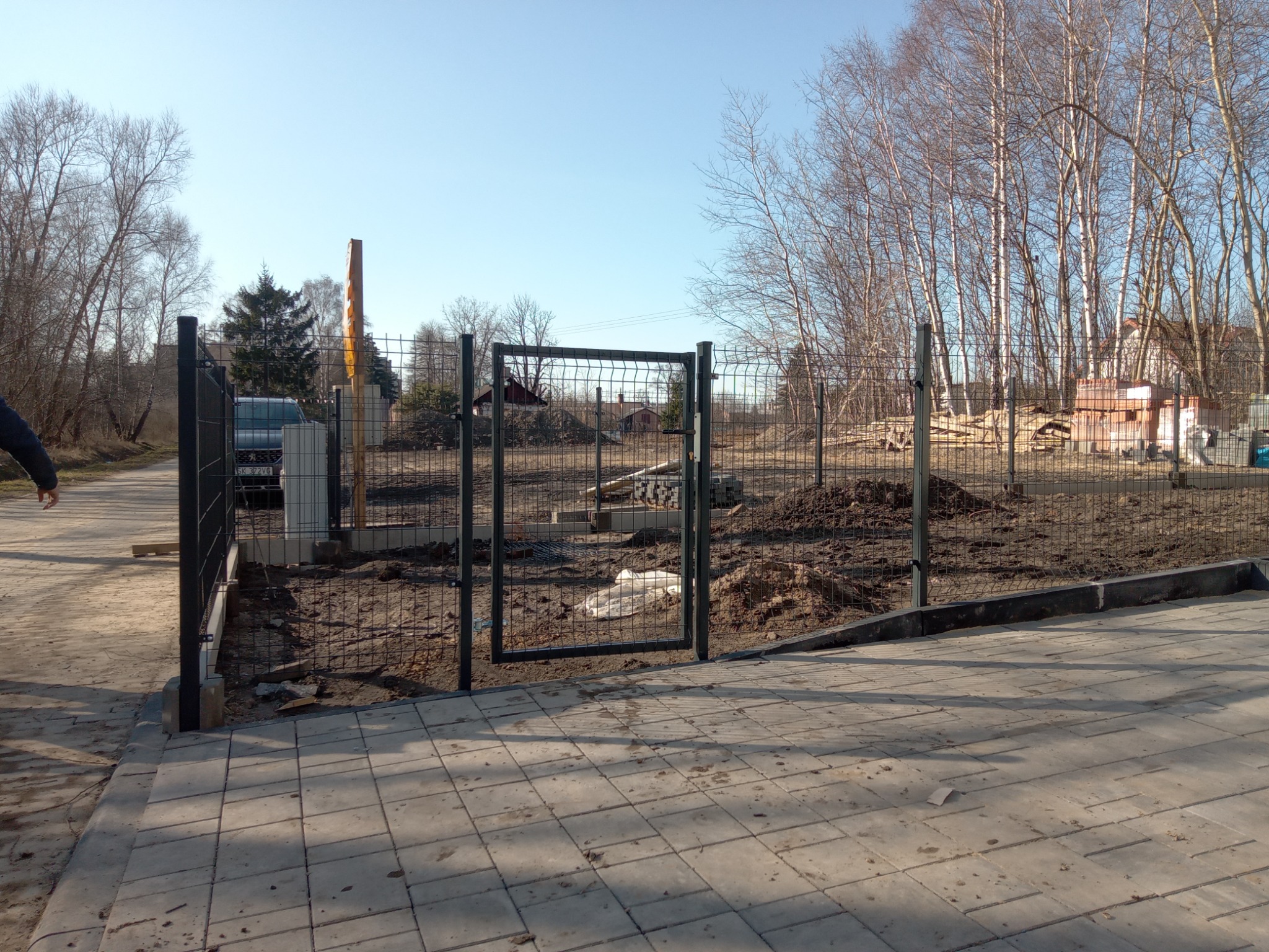 Nowo zainstalowane, czarne, panelowe ogrodzenie z bramką wejściową na posesję, widok z zewnątrz na nierówny teren z hałdami ziemi i częściowo przygotowanym podłożem pod trawnik, w tle drzewa bez...