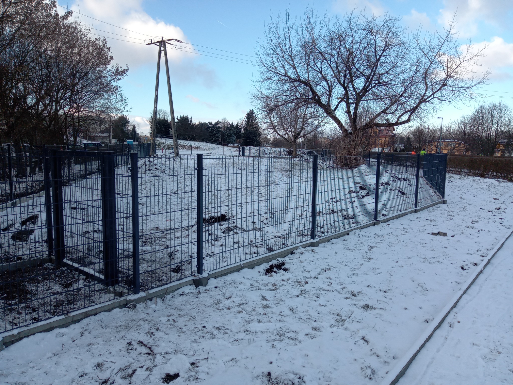 Nowo zamontowane, metalowe ogrodzenie panelowe w kolorze grafitowym na betonowej podmurówce, otaczające zasypany śniegiem teren z widocznymi śladami budowy, słup energetyczny i drzewa w tle.