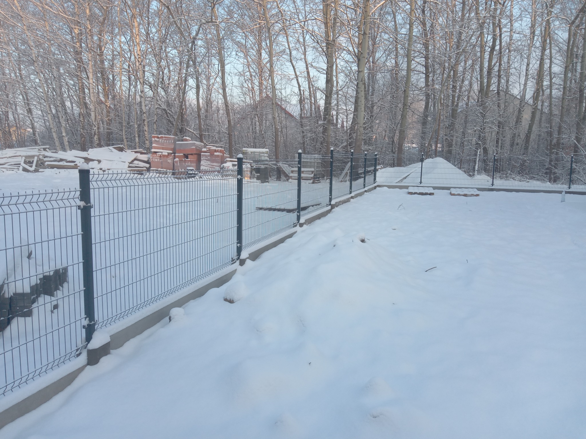 Nowo postawione, panelowe ogrodzenie w zimowej scenerii. Na śniegu widoczne ślady, w tle stos cegieł i drzewa pokryte szronem.