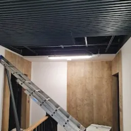 Wnętrze klatki schodowej w trakcie prac wykończeniowych, widoczna drabina aluminiowa oparta o ścianę, fragment sufitu z oświetleniem oraz okładzina ścienna imitująca kamień.