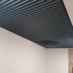 Sufit podwieszany z metalowych paneli w kolorze grafitowym, z widoczną kratką wentylacyjną w jednym z paneli.