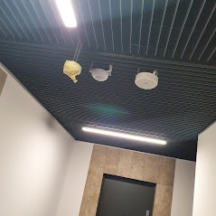 Sufit rastrowy z widocznymi elementami instalacji oświetleniowej w trakcie montażu, fragment ściany z okładziną imitującą kamień i drzwiami.