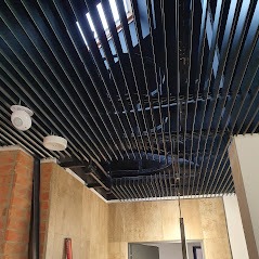 Nowoczesny sufit z metalowych listew w kolorze grafitowym, z widocznymi elementami oświetlenia i fragmentami ścian w odcieniach beżu i cegły, tworzący industrialny charakter wnętrza.