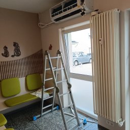 KajMar Arkadiusz Rutkowski - Wnętrze biura z zamontowaną klimatyzacją typu split, drabina oparta o ścianę, widoczny grzejnik żeberkowy, dekoracje ścienne w postaci sylwetek kotów i królika.
